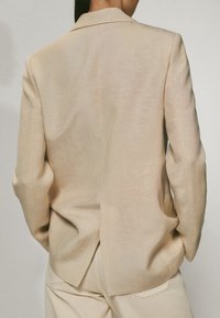Beige Blazer mit strukturiertem Design, ausgestattet mit einem Schlitz hinten, langen Ärmeln und einer subtilen Textur.