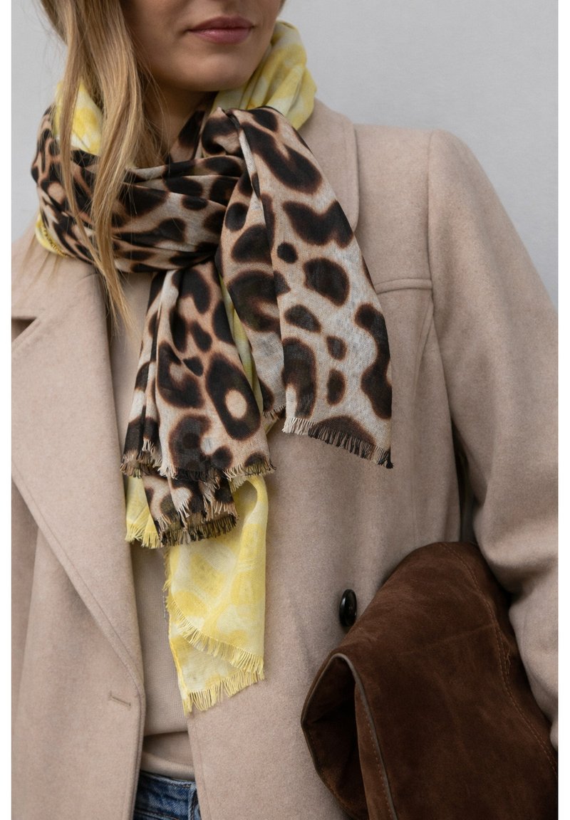Femme portant un manteau beige et deux écharpes superposées — jaune et imprimé léopard — tenant une grande pochette en suède marron sur un fond uni.