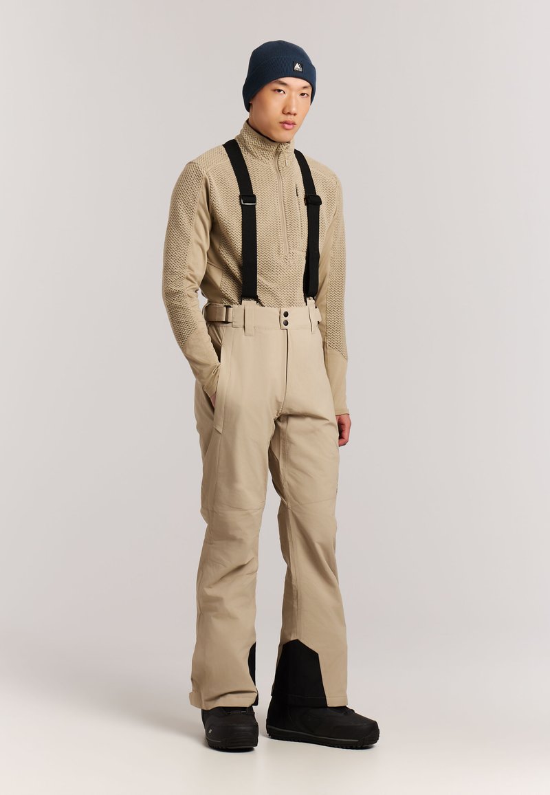 Jeune homme portant un pull zippé texturé beige, un pantalon de ski beige avec des bretelles noires et des poignets renforcés, des bottes noires et un bonnet bleu marine.