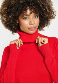 Maglione rosso a collo alto con texture a coste; maniche lunghe con polsini a coste; il tessuto appare morbido e aderente attorno al collo.