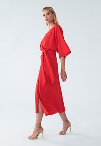 Rotes Wickelkleid mit weiten Ärmeln, tailliertem Bund und hohem Schlitz. Kombiniert mit goldenen Sandalen mit Riemchen. Glattes Material und knöchellange Passform.