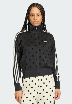 Donna con capelli intrecciati che indossa una giacca nera a pois con zip, strisce bianche e pantaloni bianchi a pois, in piedi contro uno sfondo chiaro.