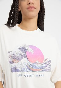 T-shirt blanc arborant un design graphique de "La Grande Vague", avec des accents violets et roses représentant des vagues et un soleil circulaire.