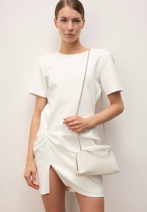 Mujer con un vestido blanco de manga corta y una abertura frontal, sosteniendo un pequeño bolso blanco con una correa de cadena plateada sobre el hombro.