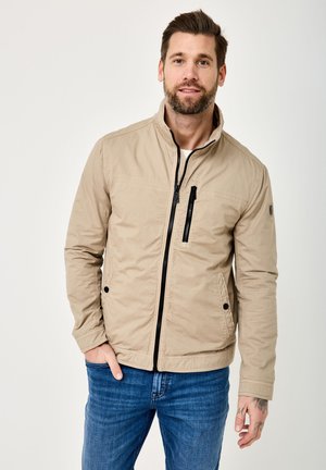 Bearded man wearing a beige zip-up jacket and blue jeans, standing with one hand in pocket against a plain light background.