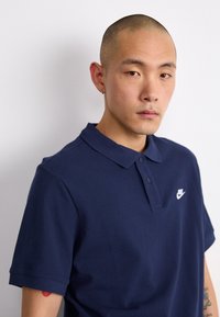 Námornícky modrá polo košeľa vyrobená z textúrovanej bavlny, s trojbodovým zapínaním a malým bielym logom Nike na hrudi.