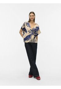 Lichtbeige blouse met een blauw tropisch bladerenprint, V-hals en losse mouwen, gecombineerd met donkere blauwe wijde jeans en rode platformsandalen.