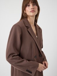 Καφέ διπλόσαρκο σακάκι από λείο ύφασμα. Διαθέτει μεγάλες πέτσες και μαύρα κουμπιά. Κομμένο κατ' ευθείαν, με υφή τελείωμα.