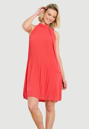 Žena sa usmieva, má krátke blond vlasy, nosí koralové plisované šaty bez rukávov s detailom na krku a stojí pred svetlým pozadím.