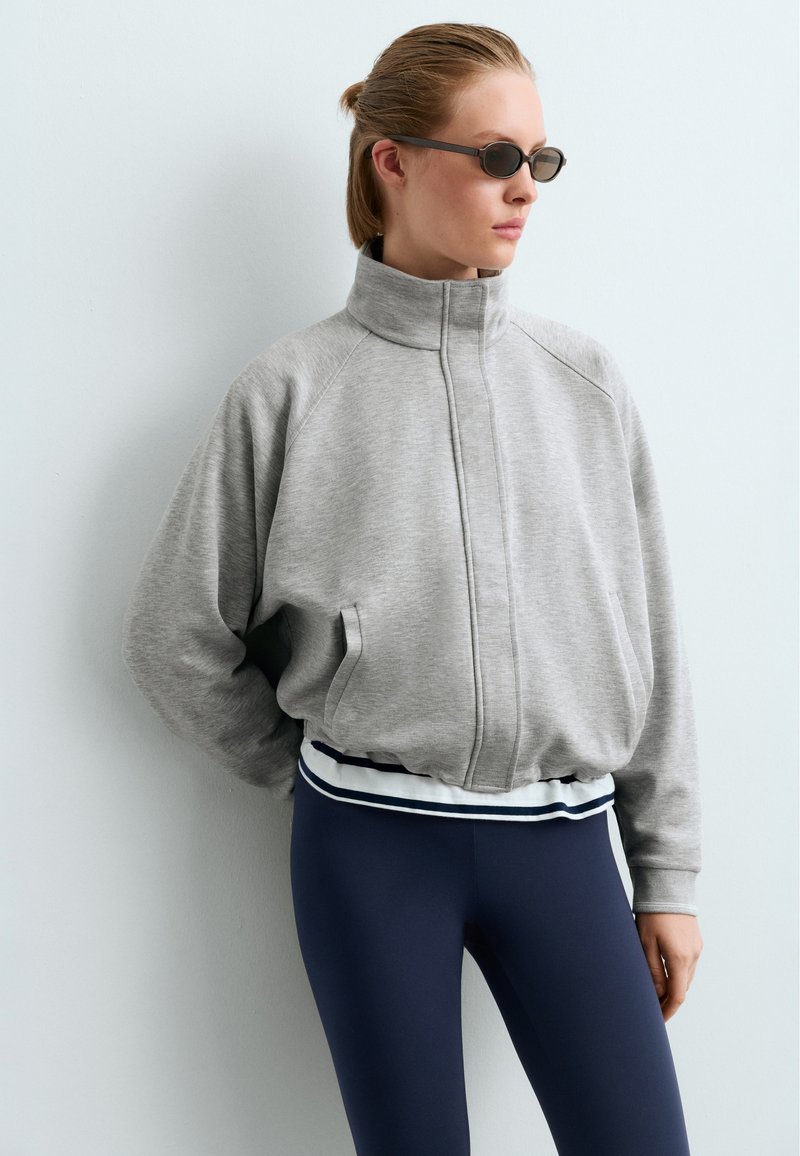 Jeune femme portant de petites lunettes de soleil noires, une veste zippée gris clair et des leggings bleu foncé, debout contre un mur blanc.