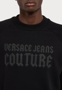 Černá mikina se žebrovaným límcem, obsahující vyvýšené černé písmo: "VERSACE JEANS COUTURE", což přidává texturu a kontrast k látce.