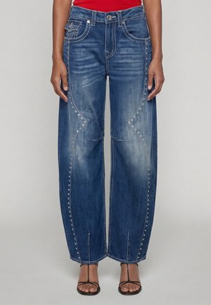 Vrouw draagt blauwe jeans met wijde pijpen en decoratieve zilveren studs langs de naden, gecombineerd met zwarte sandalen met bandjes op een effen achtergrond.