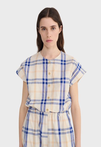 Femme portant une chemise blanche à manches courtes, boutonnée, avec un motif à carreaux bleu et orange, et un pantalon taille haute assorti, sur un fond uni.