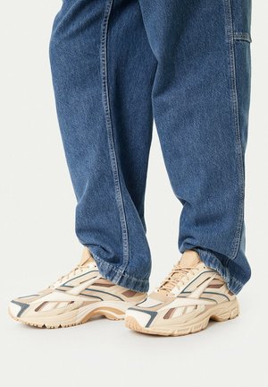 Person trägt beige-weiße Sportschuhe mit blauen Akzenten und lockere blaue Jeans vor einfarbigem Hintergrund.