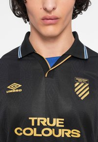 Κοντινό πλάνο του κορμού που φορά μαύρη φανέλα της Manchester Way F.C. με χρυσά λογότυπα, κείμενο "TRUE COLOURS" και κολάρο πάνω από μπλε πουκάμισο.