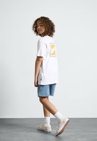 T-shirt en coton blanc avec un graphique coloré au dos, associé à un short bleu et des baskets beiges, complété par des chaussettes blanches et des semelles rugueuses.