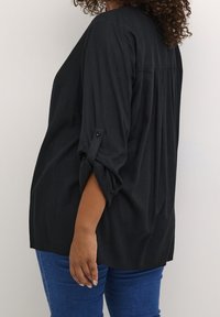 Tunique noire à coupe ample, manches longues retroussées. Présente une texture subtile et un design de découpe au dos, accompagnée d'un jean bleu.