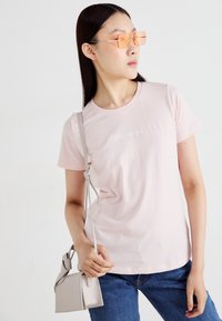 T-shirt de algodão rosa claro com mangas curtas e decote redondo, apresentando um logótipo bordado em branco. Combinado com jeans azul escuro e uma pequena bolsa cinza.