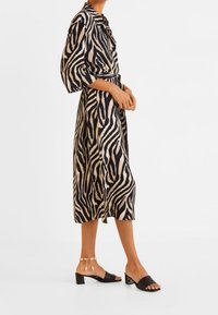 Sukienka w paski zebra w kolorze beżowym i czarnym, do połowy uda, z kołnierzykiem koszulowym i paskiem w talii. Czarne sandały na słupku z paskiem wokół kostki.