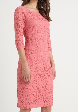 Femme portant une robe en dentelle rose mi-longue avec des manches trois-quarts et des motifs floraux, debout devant un fond blanc uni.