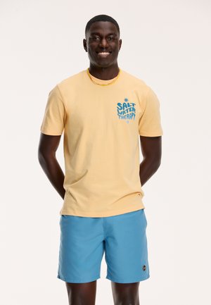 Gelbes Baumwoll-T-Shirt mit "SALT WATER THERAPY" Grafik; hellblaue Shorts mit Logo-Patch; glatte Texturen, lässige Passform.