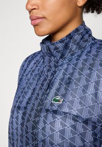 Navy patterned jacket featuring a geometric design, high collar, and a small green embroidered logo on the front. Smooth texture.