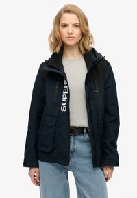 Granatowa, wodoodporna kurtka z czarnymi akcentami, z kieszeniami na zamek, logo na przodzie oraz kapturem; cechuje się luźnym krojem i trwałym materiałem.