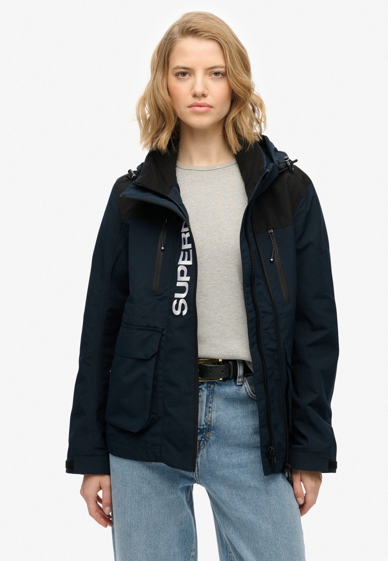 Granatowa, wodoodporna kurtka z czarnymi akcentami, z kieszeniami na zamek, logo na przodzie oraz kapturem; cechuje się luźnym krojem i trwałym materiałem.