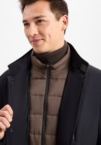 Schwarze Außenjacke mit Kragen über einer braunen Steppweste. Die Weste hat eine gesteppte Textur und einen Reißverschluss, darunter befindet sich ein brauner Rippstrickrollkragen.