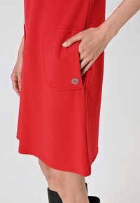 Abito rosso realizzato in un tessuto morbido con una tasca frontale. Caratterizzato da un piccolo logo decorativo sulla tasca. Design di lunghezza fino al ginocchio.