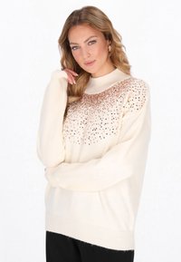 Blonde vrouw in een crème trui met gouden pailletten op de borst, poseert met één hand dichtbij haar gezicht en los haar over haar schouders.