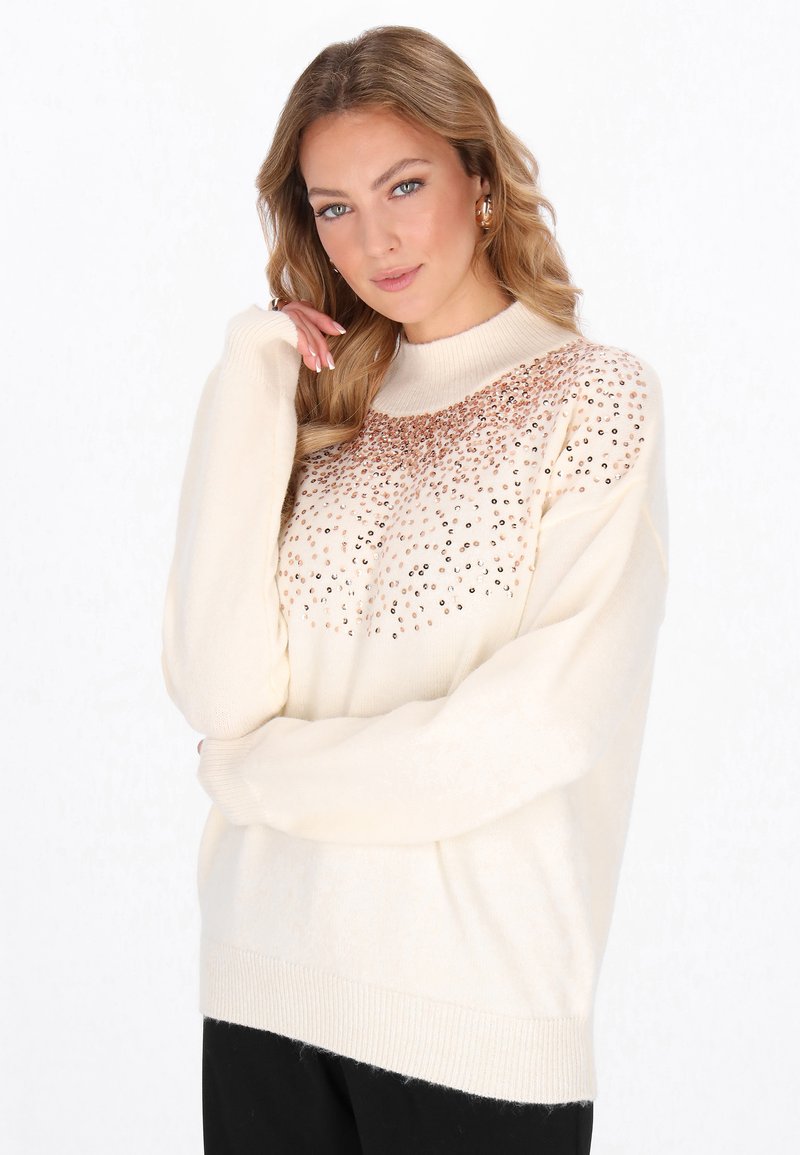 Blonde vrouw in een crème trui met gouden pailletten op de borst, poseert met één hand dichtbij haar gezicht en los haar over haar schouders.