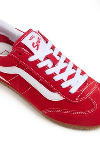 Sneaker in suede rosso con accenti bianchi, caratterizzato da una tomaia testurizzata, lacci bianchi piatti e una suola in gomma di tonalità marrone chiaro.