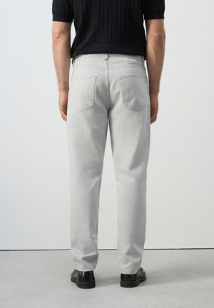 Mężczyzna w jasnoszarych dżinsach, czarnych butach i czarnej koszulce z krótkim rękawem, stojący twarzą do gładkiej białej ściany.