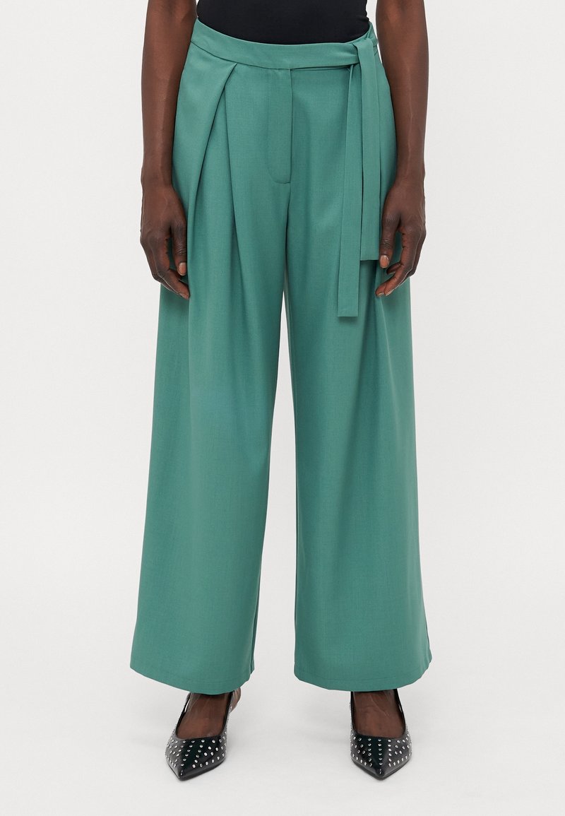 Personne grande vêtue d'un pantalon vert taille haute et à jambes larges avec un détail de ceinture, portant des chaussures noires à bout pointu avec clous, se tenant devant un fond neutre.
