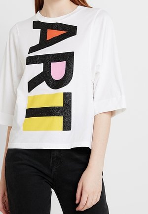 Biała koszula z długim rękawem z dużymi czarnymi literami tworzącymi słowo „ART”, wypełnionymi pomarańczowymi, różowymi i żółtymi kształtami, noszona z czarnymi spodniami.