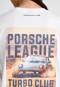 Hvid t-shirt med et stort grafisk motiv af en sort bil, der kaster støv op, med "PORSCHE LEAGUE TURBO CLUB" trykt i fed, slidt tekst.