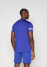 Μπλε αθλητικό μπλουζάκι με κοντά μανίκια, με στρογγυλή λαιμόκοψη και λευκό εκτυπωμένο λογότυπο στο αριστερό μανίκι, συνδυασμένο με μπλε σορτς.