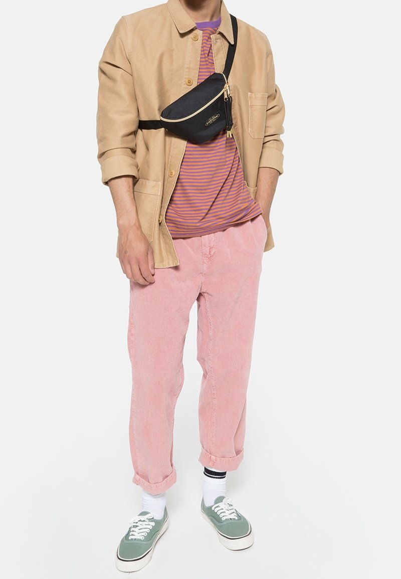 Homme portant une veste beige, une chemise rayée violette, un pantalon rose, des baskets vertes, des chaussettes blanches et un sac bandoulière noir sur fond blanc.