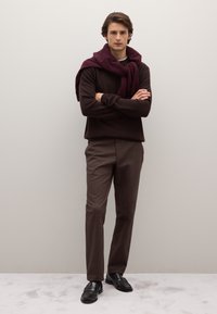 Maglione marrone scuro indossato sopra una camicia bianca, abbinato a pantaloni marrone chiaro e mocassini neri. Il modello ha una sciarpa bordeaux appesa attorno al collo.
