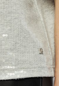 Hallikas särahelkuritega üleminekupluus, millel on tekstuuriga kootud kangas, väike hõbedane logo silt ja äär, millel on kaunistused.