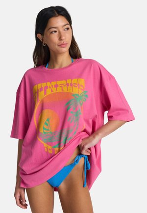 Oversized lyserød bomulds-T-shirt med "SUNRISE" grafik i gul og grøn, palmer og et solnedgangsdesign; kombineret med blå bikini med bindebånd i siderne.