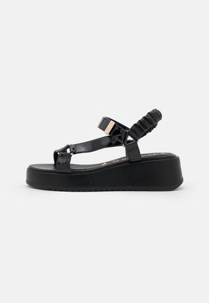 Black patent leather wedge sandals with a thick black platform, crisscross straps, and a gathered ankle strap detail. Gold-tone hardware accents.