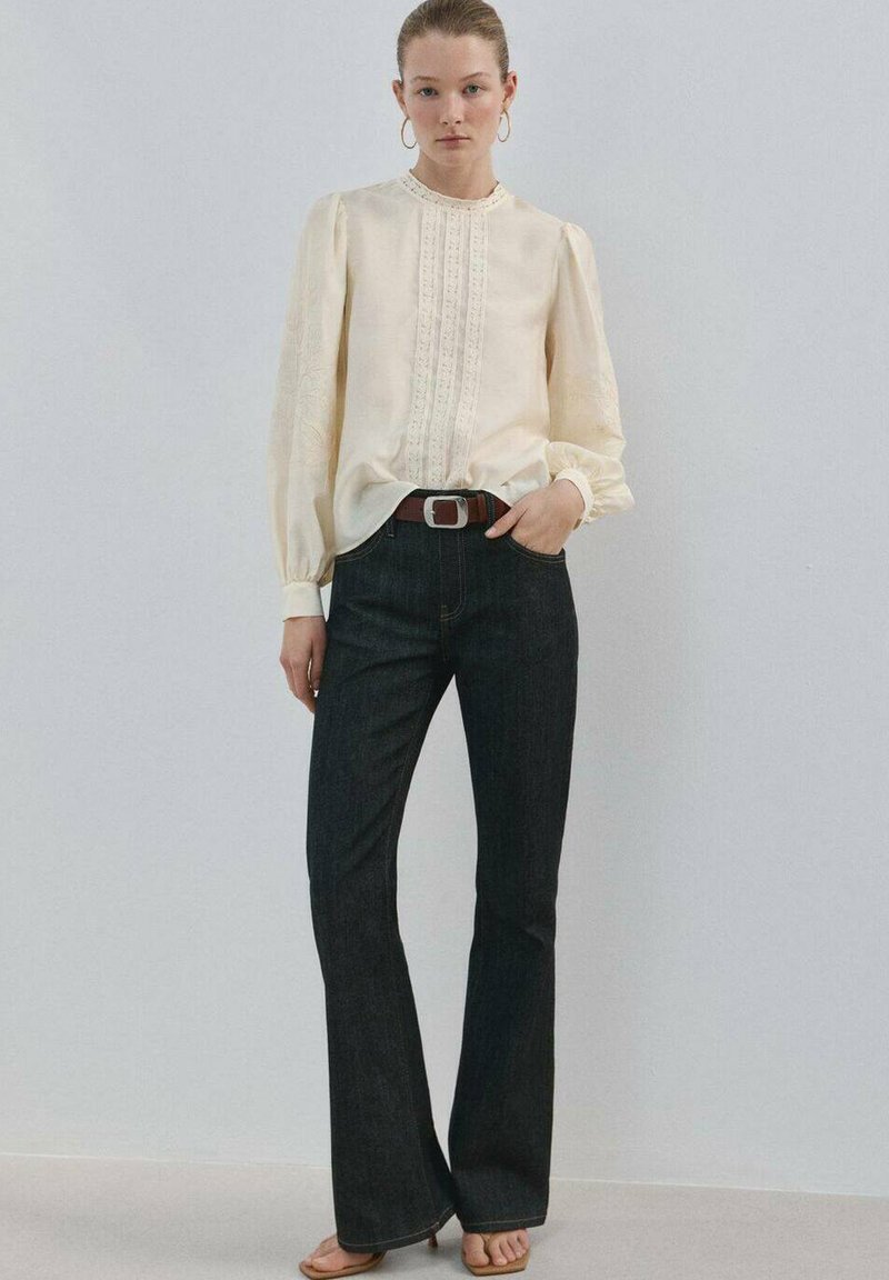 Junge Frau, die eine cremefarbene Bluse mit Spitzeneinsätzen und Puffärmeln, dunkle ausgestellte Jeans, braunen Gürtel und offene High Heels trägt, steht vor einer schlichten Wand.