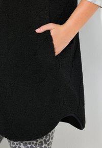 Käsi pistetty musta tekstuuri fleece riide küljetaskusse, näidates hall leopardimustriga legginsite osa ja valget varrukat.