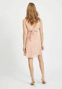 Žena s vlnitými vlasy, která má na sobě bezramínkové, světle růžové šaty ke kolenům s detailem na zavazování na zádech a béžové sandály na vysokém podpatku.