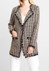 Texturerad kavaj i svart, beige och krämfärgad; har fransdetaljer, slagkrage och två framfickor. Knappar nära midjan.