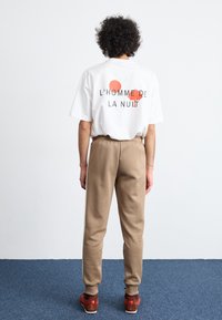 Camiseta blanca con estampado gráfico que presenta círculos rojos y el texto "L'Homme de la Nuit." Pantalones deportivos beige y zapatillas rojo-marrones.