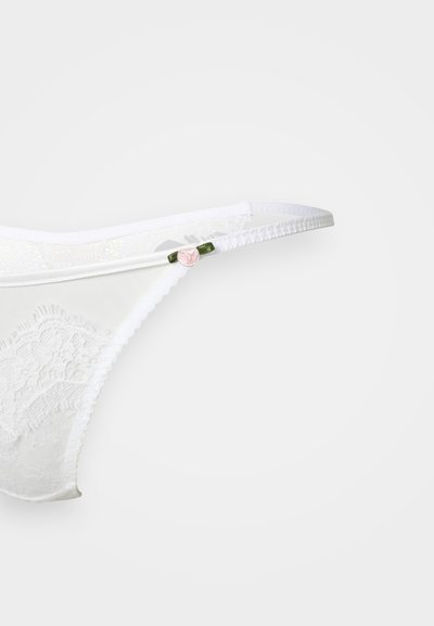 String en dentelle blanche avec un motif floral, des straps élastiques fins, et un petit accent en rose pâle sur une boucle verte. Texture transparente et délicate.