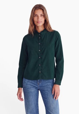 Kvinde med langt hår iført en mørkegrøn bluse med knapper og flæser og blå jeans, stående mod en ensfarvet hvid baggrund.